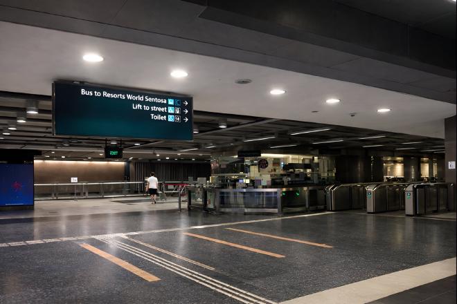An underground MRT station concourse in Singapore with ticket gates, a directional sign pointing to Resorts World Sentosa, and a pedestrian.
